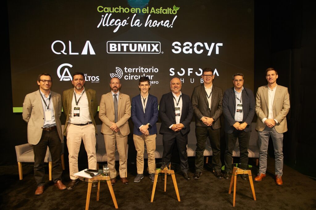 Grupo de personas posando en un escenario frente a un fondo con el mensaje “Caucho en el Asfalto – ¡Llegó la hora!” y logotipos de empresas y organizaciones relacionadas con economía circular e infraestructura.