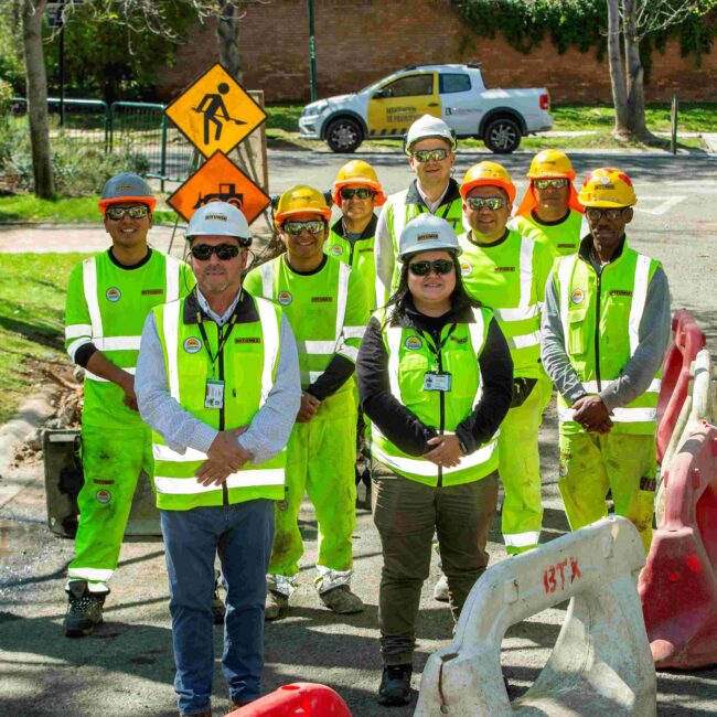 Grupo de personal de obras urbanas con chalecos reflectantes y cascos de seguridad, de pie en una calle delimitada por barreras y señalización vial.