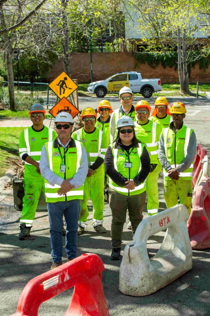 Grupo de personal de obras urbanas con chalecos reflectantes y cascos de seguridad, de pie en una calle delimitada por barreras y señalización vial.