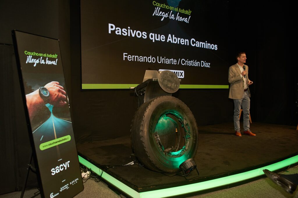 Persona exponiendo en escenario frente a una presentación titulada “Pasivos que Abren Caminos”, con un neumático reciclado exhibido como ejemplo de reutilización de caucho en pavimentos.