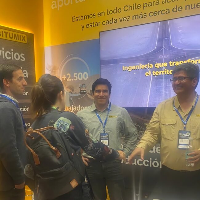Equipo de Bitumix explica sus servicios a visitantes en stand de Exponor 2026, frente a paneles informativos de minería y pantalla con imagen aérea de carretera.