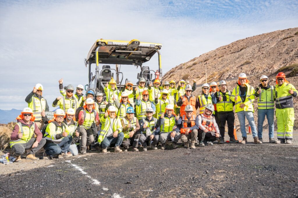 Grupo de trabajadores con elementos de protección personal posando frente a una máquina finisher sobre una carretera en construcción, en una zona montañosa y desértica.