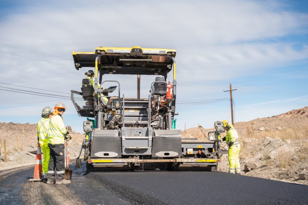 Finisher de pavimentación extendiendo mezcla asfáltica sobre una carretera, con trabajadores equipados con elementos de seguridad operando y supervisando las labores en un entorno desértico.