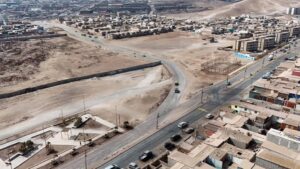 Fotografía aérea de Alto Hospicio que muestra una vía en construcción conectada a una avenida existente, terrenos intervenidos para obras urbanas, un pequeño parque en primer plano, viviendas y edificios residenciales alrededor, con tránsito vehicular y paisaje desértico característico de la zona.