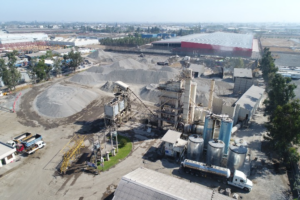 Vista aérea de una planta industrial con equipos de procesamiento, estanques metálicos, camiones y grandes pilas de áridos almacenadas en un recinto de operaciones industriales.