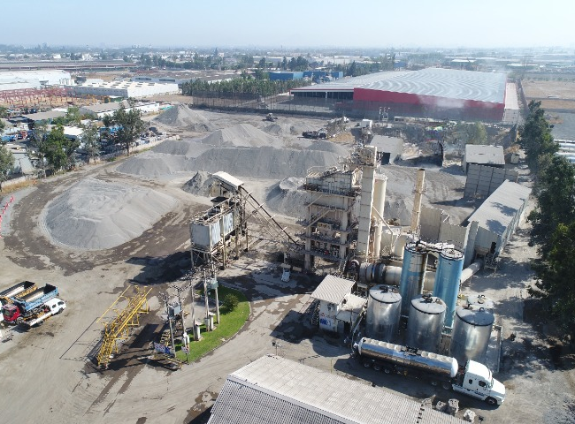 Vista aérea de una planta industrial con equipos de procesamiento, estanques metálicos, camiones y grandes pilas de áridos almacenadas en un recinto de operaciones industriales.