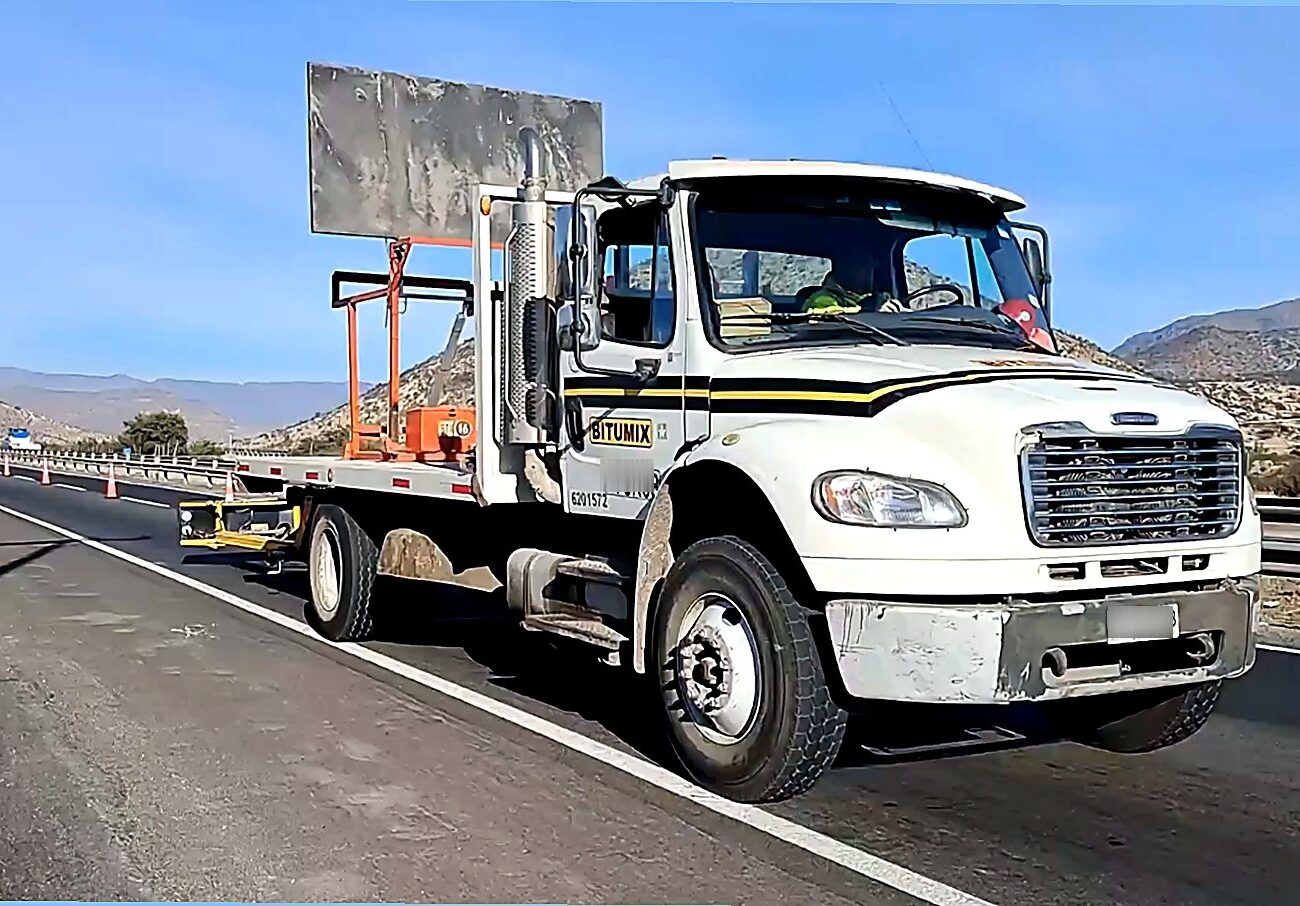 Camión de señalización vial estacionado en el borde de una carretera con conos de tráfico, barreras de contención y un panel de señalización montado en la plataforma trasera, en un entorno montañoso y despejado.
